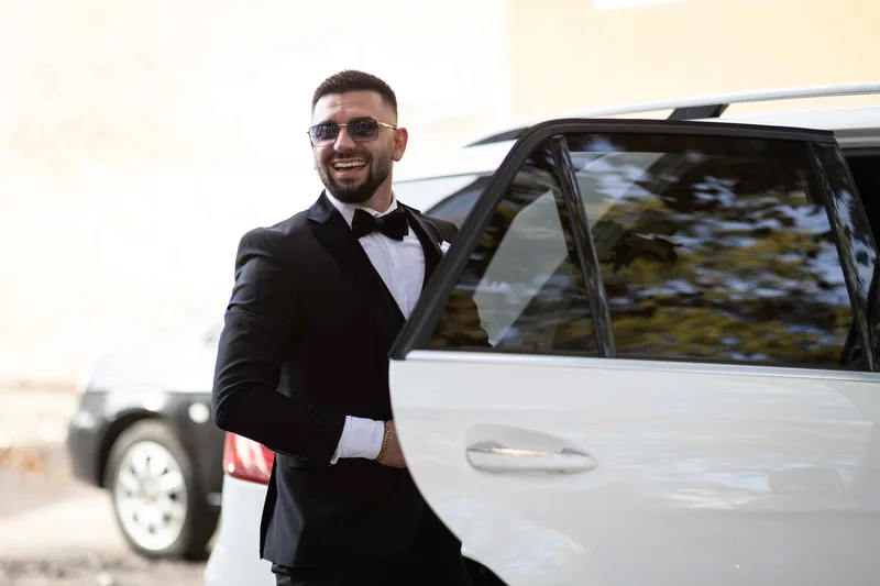 Усмихнат младоженец в смокинг с папийонка до бял автомобил